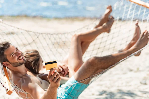 Pareja joven sosteniendo la tarjeta de crédito de oro y tumbado en la hamaca en la playa cerca del mar - foto de stock