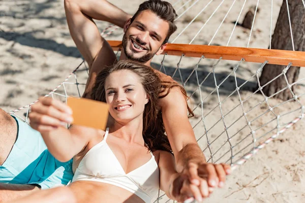 Счастливая красивая пара, держащая золотую кредитную карту и расслабляясь на гамаке на пляже — стоковое фото
