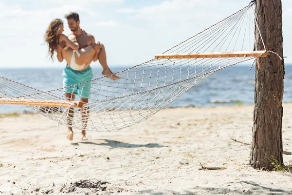 Foco selectivo del hombre que sostiene a novia en las manos, con la hamaca en la playa en primer plano - foto de stock