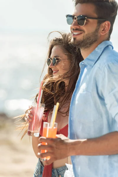 Belo casal em óculos de sol segurando coquetéis e olhando para o mar — Fotografia de Stock