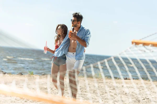 Casal com coquetéis de álcool andando na praia perto da rede — Fotografia de Stock