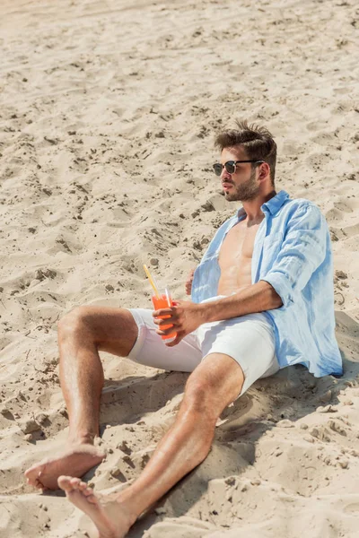 Homem barbudo bonito em óculos de sol sentado na praia de areia com copo de coquetel de álcool — Fotografia de Stock
