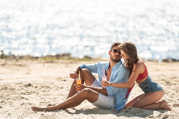 Pareja con cócteles sentados en la playa cerca del mar en verano — Stock Photo