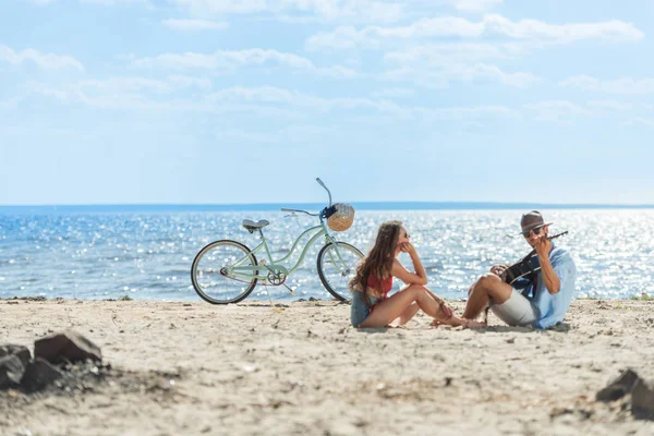 Handsome man playing on guitar for his girlfriend and resting on beach with bike — Stock Photo