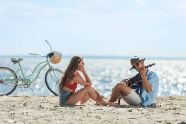 Hombre tocando la guitarra acústica para su novia y sentado en la playa con bicicleta - foto de stock