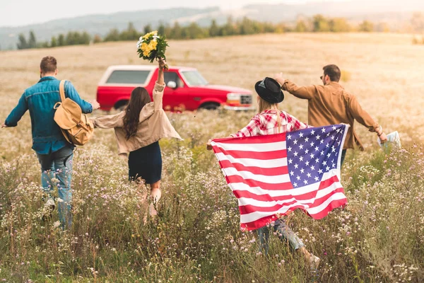 Задний вид группы молодых американских путешественников с флагом прогуливаясь по цветочному полю во время автомобильной поездки — стоковое фото
