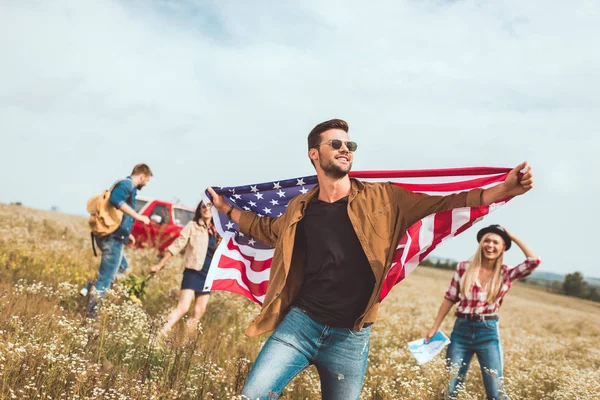 Joven guapo sosteniendo bandera de EE.UU. y caminando por el campo con amigos durante el viaje - foto de stock
