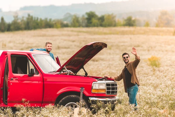 Jóvenes de pie cerca del coche con el motor roto en el campo y saludando a la cámara — Stock Photo