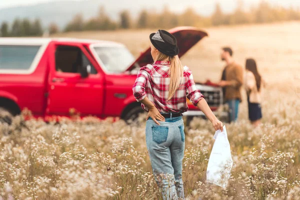 Vista posteriore di giovane donna con mappa a piedi in auto rotta nel campo di fiori mentre i suoi amici in piedi su sfondo — Foto stock