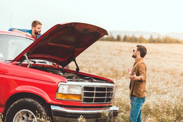 Young men standing near car with broken engine in field — Stock Photo