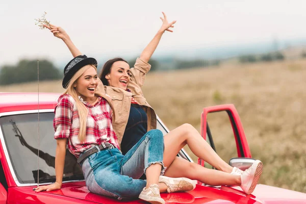 Happy young girlfriends sitting on car hood with raised hands and showing peace signs — Stock Photo
