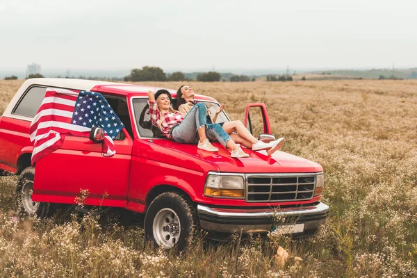 Beautiful young girlfriends relaxing on car hood in field — Stock Photo