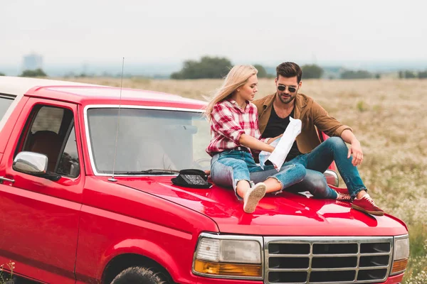 Travelling young couple sitting on car engine hood and navigating with map — Stock Photo