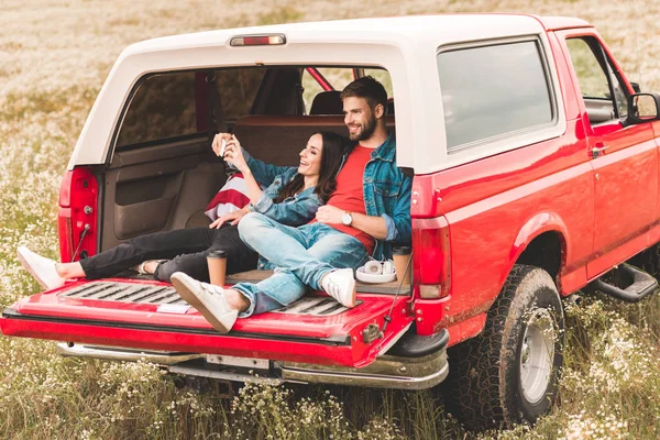 Hermosa pareja joven tomando selfie con teléfono inteligente mientras se relaja en el maletero del coche - foto de stock