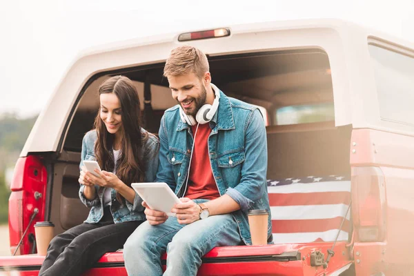 Glückliches junges Paar benutzt Gadgets, während es während der Fahrt im Kofferraum sitzt — Stockfoto