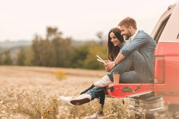 Vista laterale di felice giovane coppia utilizzando gadget mentre seduto nel bagagliaio dell'auto durante il viaggio — Foto stock