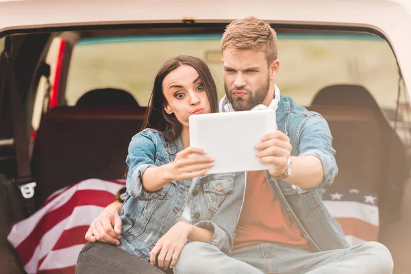 Beau jeune couple faisant selfie avec tablette tout en étant assis dans le coffre de la voiture pendant le voyage — Photo de stock