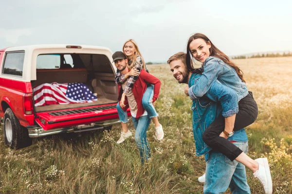 Young happy women piggybacking on boyfriends in flower field during car trip — Stock Photo