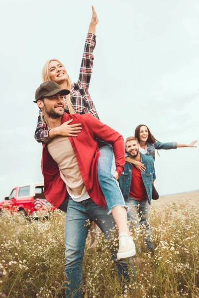 Mulheres jovens piggybacking em namorados no campo de flores — Fotografia de Stock