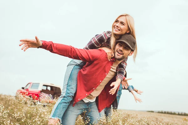 Sonriente joven mujer piggybacking en novio en flor campo con borrosa coche en el fondo - foto de stock