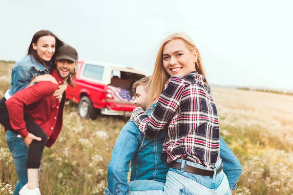 Les jeunes femmes piggyback sur copains et regarder la caméra dans le champ — Photo de stock