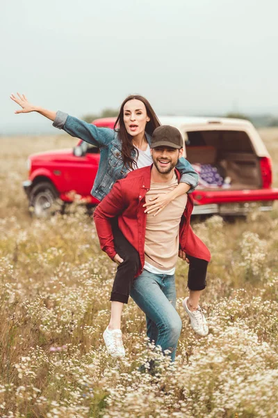 Heureux jeune femme piggybackking sur copain dans champ de fleurs avec voiture floue sur fond — Photo de stock