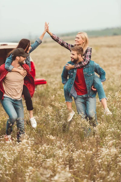 Jóvenes mujeres piggybacking en novios y dando alta cinco en el campo de flores - foto de stock