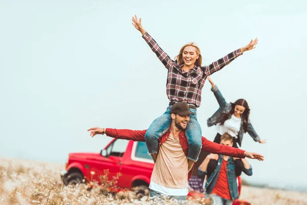 Mulheres jovens montando em namorados ombros e levantando as mãos no campo durante a viagem de carro — Fotografia de Stock