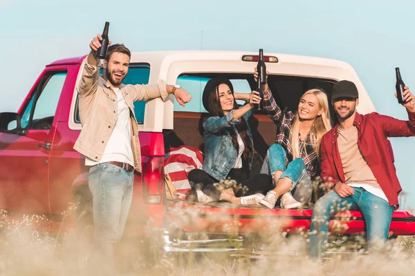 Group of young people toasting with bottles of beer while sitting in car trunk — Stock Photo