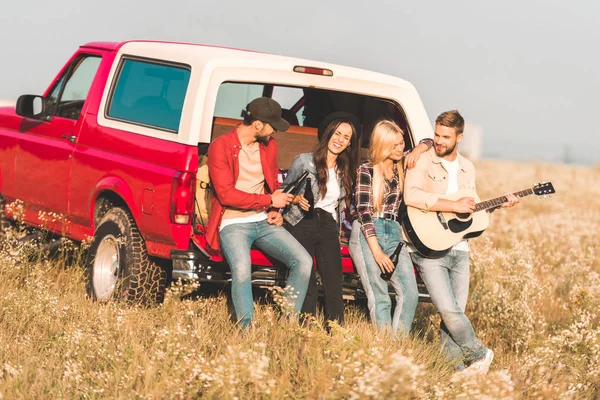 Gruppe fröhlicher junger Leute trinkt Bier und spielt Gitarre, während sie es sich im Kofferraum eines Autos auf einem Blumenfeld gemütlich macht — Stockfoto