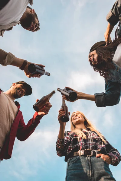 Vista inferior del grupo de jóvenes felices tintineando botellas de cerveza frente al cielo nublado - foto de stock