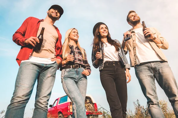 Bottom view of group of happy young people with beer bottles and guitar on field — Stock Photo