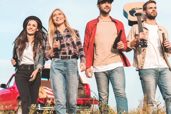 Bottom view of group of young fiends with beer bottles and guitar walking by field — Stock Photo