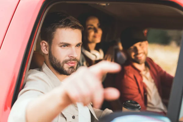 Handsome young man pointing somewhere while riding car with friends — Stock Photo