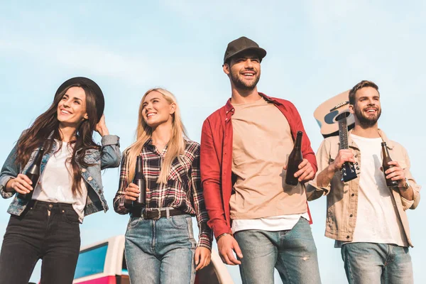 Bottom view of group of happy young people with beer bottles and guitar walking by field — Stock Photo