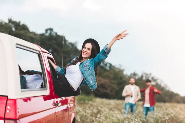Young woman outstretching from car window and raising hand while men standing blurred on background in flower field — Stock Photo