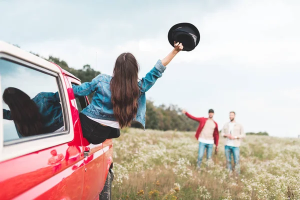 Young woman outstretching from car window and greeting to men standing in flower field — Stock Photo