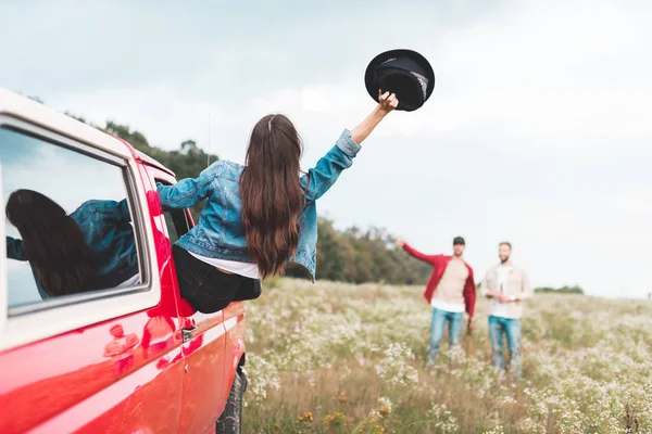 Junge Frau streckt sich aus Autoscheibe und grüßt Männer, die im Blumenfeld stehen — Stockfoto