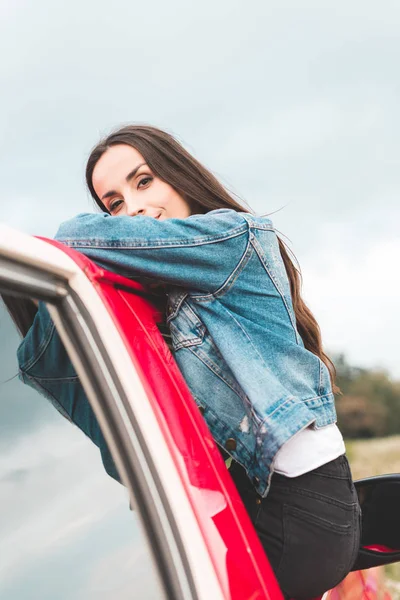 Bela jovem mulher inclinada no veículo vermelho ao ar livre e olhando para a câmera — Fotografia de Stock