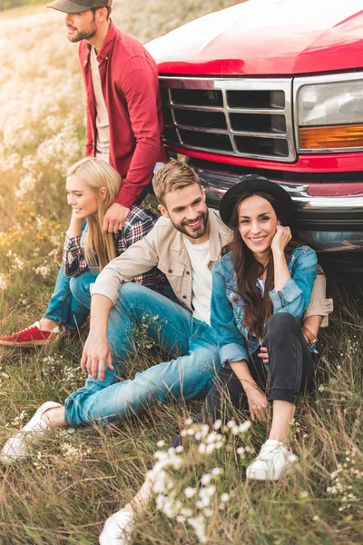 Group of happy young car travellers sitting on flower field and leaning back on vintage truck — Stock Photo