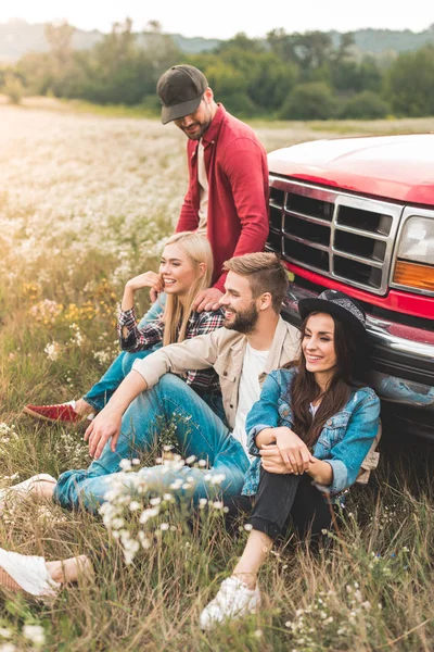 Group of young car travellers sitting on flower field and leaning back on vintage truck — Stock Photo