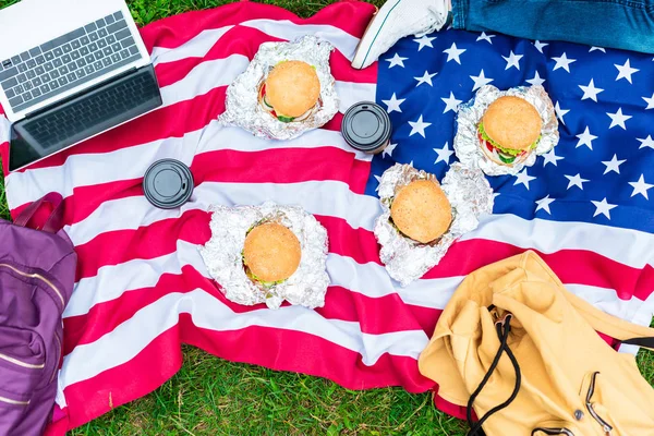 Vista parcial del hombre acostado cerca de la bandera americana con el ordenador portátil, hamburguesas y café para ir sobre hierba verde - foto de stock