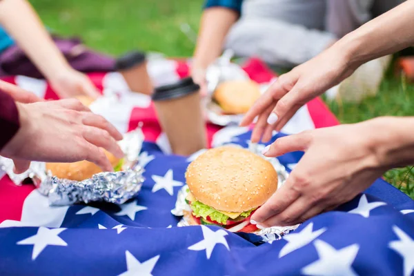 Vista parcial de amigos con hamburguesas y bandera americana sobre hierba verde en parque - foto de stock