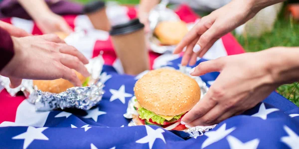 Частичный вид друзей с гамбургерами и американским флагом на зеленой траве в парке — стоковое фото