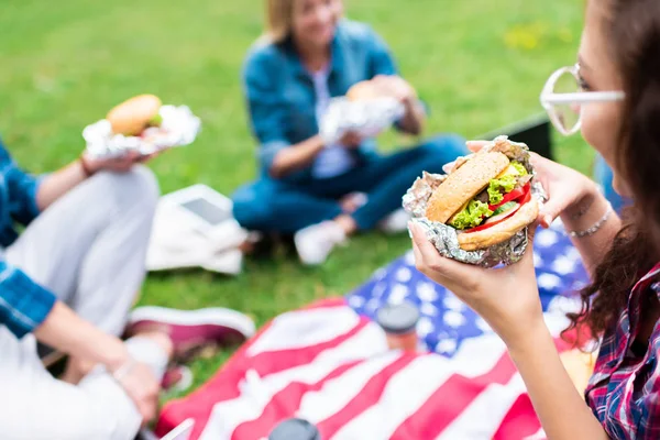 Частичный вид друзей с гамбургерами и американским флагом на зеленой траве в парке — стоковое фото