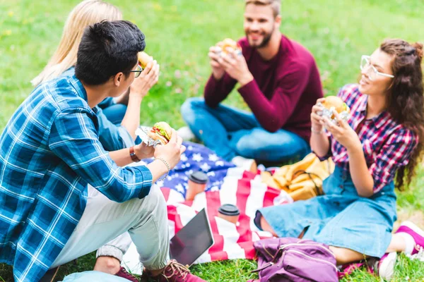 Multiracial students with burgers and american flag resting in park — Stock Photo