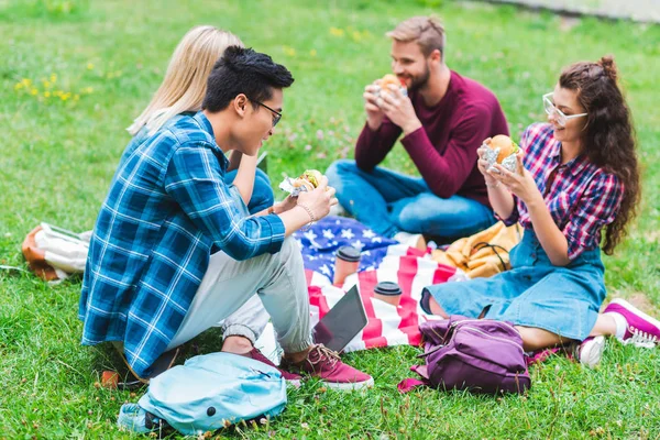 Estudiantes multirraciales con hamburguesas y bandera americana descansando en el parque - foto de stock