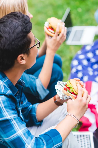 Enfoque selectivo de los estudiantes con hamburguesas sentados en hierba verde con bandera americana - foto de stock