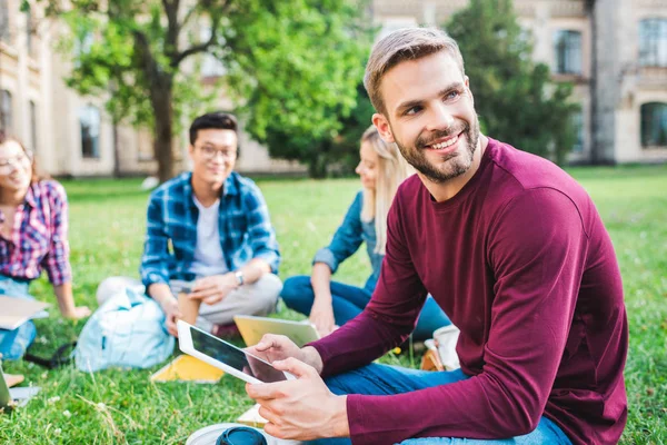 Studenti sorridenti multirazziali con dispositivi digitali seduti su erba verde nel parco — Foto stock