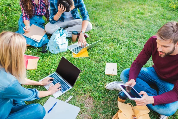 Vista parcial de estudiantes multiétnicos con cuadernos y dispositivos digitales sentados sobre hierba verde en el parque - foto de stock